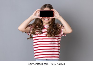 Portrait Of Little Girl Wearing Striped T-shirt Covering Eyes With Cellphone And Smiling, Unknown Child Hiding Face With Mobile Phone, Anonymous User. Indoor Studio Shot Isolated On Gray Background.