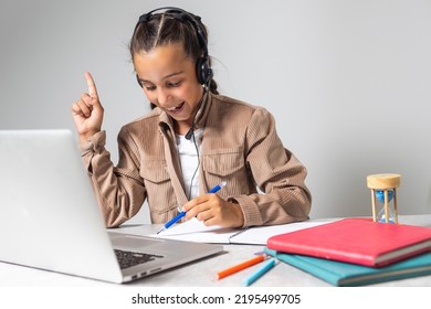 Portrait Of Little Girl In Wireless Headset Using Laptop, Studying Online At Home, Interested Happy Student Typing On Keyboard Looking At Pc Screen, Watching Webinar, Online Course, Doing Homework.