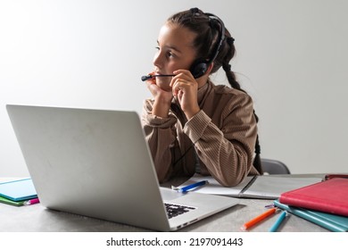 Portrait Of Little Girl In Wireless Headset Using Laptop, Studying Online At Home, Interested Happy Student Typing On Keyboard Looking At Pc Screen, Watching Webinar, Online Course, Doing Homework.