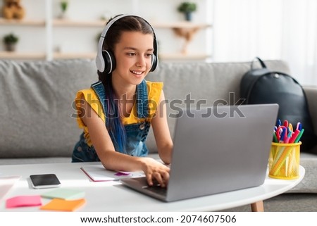 Portrait of little girl in wireless headset using laptop, studying online at home, interested happy student typing on keyboard looking at pc screen, watching webinar, online course, doing homework