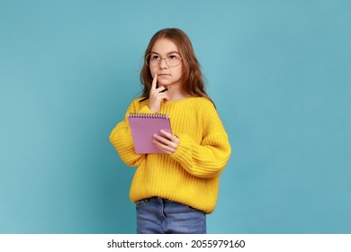 Portrait of little girl writing in notebook, thinking, taking notes, school child doing homework, wearing yellow casual style sweater. Indoor studio shot isolated on blue background.