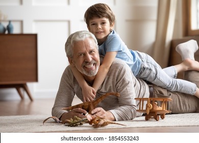 Portrait Little Grandchild Lying On Happy Mature Grandfather Back, Looking At Camera, Overjoyed Smiling Older Man And Adorable Boy Grandson Hugging, Having Fun, Lying On Warm Floor At Home