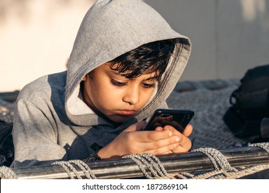 Portrait Of Little Indian School Kid Wearing Hoodie Using Mobile Phone At Home. Concept Of Online Education During Corona Virus Pandemic