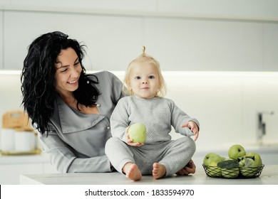 Portrait Of A Little Interesting Girl Blonde Baby Kids And A Cheerful Young Kind Mother Brunette, Playing In The Kitchen With Green Apples Show A Warm Attitude To Each Other. Banner For Advertising