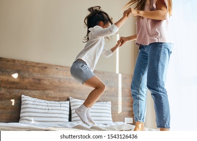 Portrait Of Little Joyful Six Years Old Girl Happily Jumping On Bed Together With Sister In Brightly Lighted Bedroom, Shot From Below, Family Joy Concept