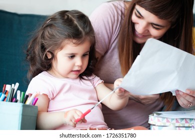 Portrait Of Little Kid Sitting On Sofa Together With Young Mother And Cutting Paper Into Small Pieces With Scissor.Self Isolated At Home.Early Childhood Development Activity, Learning And Education.