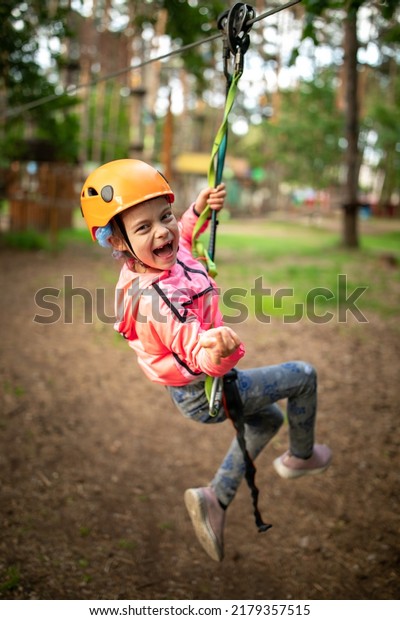 Portrait of little laughing\
girl in helmet and harness moving on trail in the sky rope park in\
summer