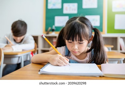 Portrait Of Little Pupil Writing At Desk In Classroom At The Elementary School. Student Girl Study Doing Test In Primary School. Children Writing Notes In Classroom. Education Knowledge Concept