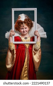 Portrait Of Little Red-headed Girl, Child In Costume Of Royal Person Holding Picture Frame On Dark Green Background. Concept Of Historical Remake, Comparison Of Eras, Medieval Fashion, Emotions, Queen