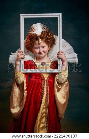 Portrait of little red-headed girl, child in costume of royal person holding picture frame on dark green background. Concept of historical remake, comparison of eras, medieval fashion, emotions, queen