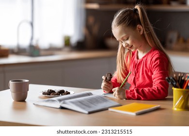 Portrait Of Little Schoolgirl Having Snacks While Doing Homework, Smiling Preteen Female Child Eating Cookies And Writing In Workbook While Sitting At Desk In Home Interior, Copy Space - Powered by Shutterstock