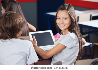 Portrait of little schoolgirl holding digital tablet at desk with classmates in classroom