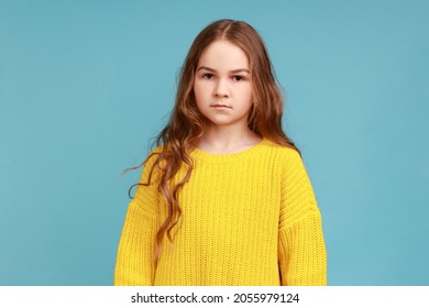 Portrait Of Little Serious Child Girl Looking At Camera With Confident Concentrated Expression, Wearing Yellow Casual Style Sweater. Indoor Studio Shot Isolated On Blue Background.