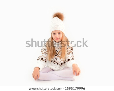 portrait of a little smiling blonde girl in a white and red  hat and scarf on a white background.Winter clothes. Cute blonde girl in white knitted hat and sweater.