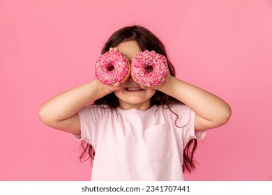 Portrait of a little smiling girl with curly hair and two appetizing donuts in her hands, closes her eyes with donuts, on a pink background, a place for text. Dieting concept and junk food.