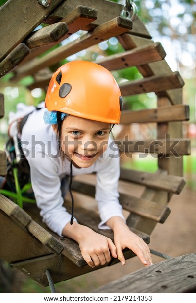 Portrait of little smiling girl in helmet\
and harness on trail in sky rope park in\
summer
