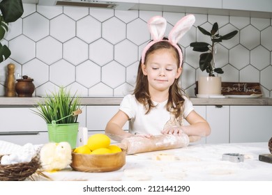 Portrait of little smiling studiously girl cooking, helping chef making easter dough cookies. Preparing dough, rolling pin with flour. Wood plate eggs and plant, chicken near child. Kitchen utensil