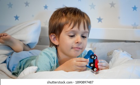 Portrait Of Little Smiling Toddler Boy Lying In Bed At Night And Playing With Two Wooden Toy Cars Before Going To Sleep