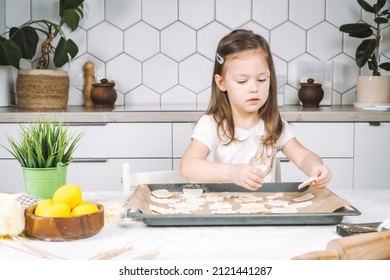 Portrait of little studiously girl chef, sitting on chair, making different shape dough cookies on dripping pan. Wooden plate of yellow easter eggs and plant pot near child. Kitchen utensil indoor.