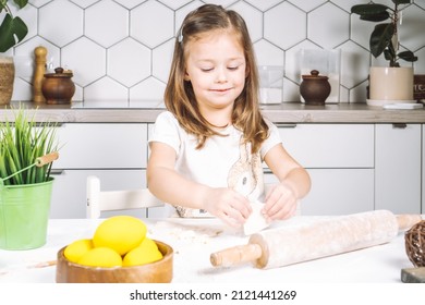 Portrait of little studiously girl sitting on chair, assistance, making different shape cookies. Preparing dough, rolling pin with flour. Sculpting shape biscuit. Plate yellow easter eggs, plant pot