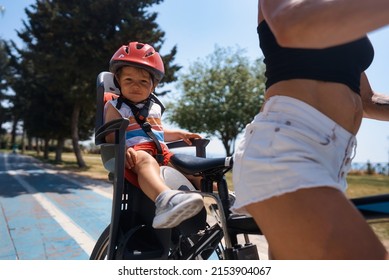 Portrait of little toddler boy with security helmet on the head sitting in bike seat and his mother with bicycle. Safe and child protection concept. Family and weekend activity trip