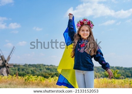 Portrait of little Ukrainian girl wearing national clothes with embroidery and wreath of flowers holding flag of Ukraine outdoor. A symbol of faith in peace for all people of Ukraine.