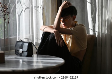 Portrait Of Lonely Asian Gay Teenager With Crop Top Shirt And Orange Glasses Sitting With Knees Up On Chair, Lifestyle Fashion LGBTQ Concept.