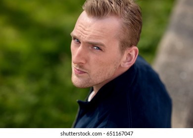 Portrait Of A Lonely Man Sitting Down Outdoors On A Bright Day With A Green Grass Field And River Behind Him. Photo Shot With Short Depth Of Field.