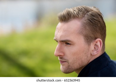 Portrait Of A Lonely Man Sitting Down Outdoors On A Bright Day With A Green Grass Field And River Behind Him. Photo Shot With Short Depth Of Field.