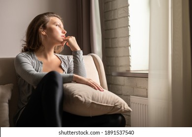 Portrait of lonely sad сaucasian young woman sitting near window of apartments. 