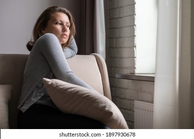 Portrait of lonely sad сaucasian young woman sitting near window of apartments. 