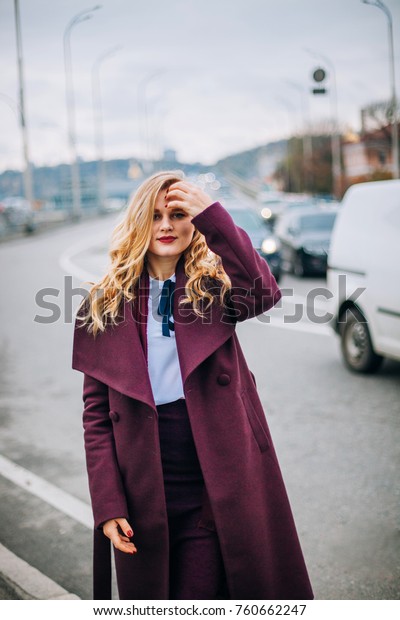 portrait of Lonely young beautiful woman girl
standing and posing on the highway on the road, cars lights, city
lights, cloudy and cold windy weather, the wind blowing long
stylish burgundy coat