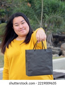 Portrait Long Black Hair Fat Asian Woman Chubby, Beautiful, Cute Wearing Yellow Blouse. Standing Smiling Happily And Freely In Park. With Hand Holding Black Paper Bag After Walking Around Shopping