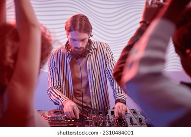 Portrait of long haired DJ by turntable in neon lights entertaining dancing crowd in nightclub - Shutterstock ID 2310484001