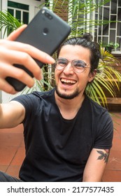 Portrait of long haired tattooed man with beard holding a phone on his hand smiling and laughing taking a selfie. green plants and brick background