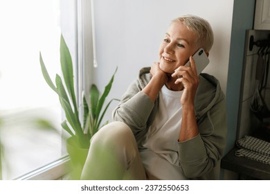 Portrait of lovely active grandma talking on phone with her grandson resting on windowsill after training outside, senior female with short hair having nice conversation on smartphone. Selective focus - Powered by Shutterstock