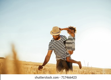 Portrait Of A Lovely African American Father Carrying His Daughter While She's Playing With His Hat In The Fields.