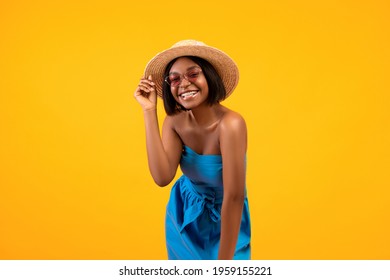 Portrait of lovely African American woman in beautiful dress, straw hat and sunglasses on orange studio background. Millennial black lady wearing trendy summer outfit, ready for beach vacation