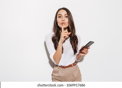 Portrait of a lovely asian businesswoman holding mobile phone and showing silence gesture isolated over white background