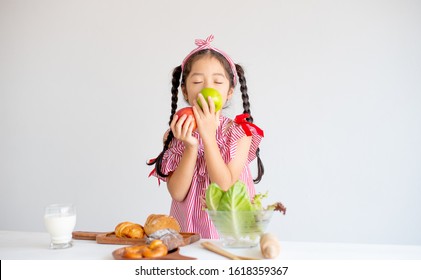 Portrait of lovely Asian little girl smell the apples with bowl of vegetables and bread on table and stand in front of white wall background. Concept of healthy food for children as fruit and milk.