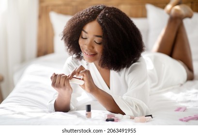 Portrait of lovely black woman filing her nails, making manicure on bed at home. Pretty African American lady in domestic robe applying decorative polish. Spa procedures, beauty treatments concept - Powered by Shutterstock