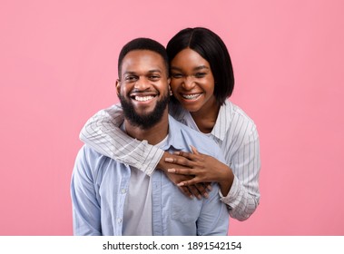 Portrait of lovely black woman hugging her partner from behind, smiling and feeling happy on pink studio background. Romantic sweethearts embracing and showing their love on Valentine's Day