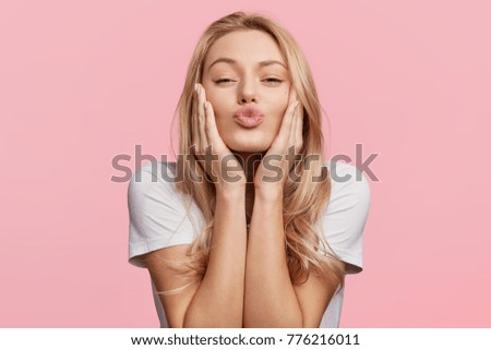 Portrait of lovely blonde female rounds lips as going to recieve kiss from boyfriend, looks at camera with pleased expression, isolated over pink background. Beautiful young woman poses in studio