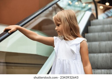Portrait of lovely blonde little girl 5-6 year old in white dress going down escalator. Photo of charming funny kid goes down, alone smiling looking away. Happy childhood concept. Copy ad text space