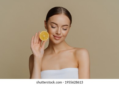 Portrait of lovely brunette young woman concentrated down holds fresh lemon slice recommends natural beauty products prefers natural organic facial treatment wrapped in bath towel poses indoor