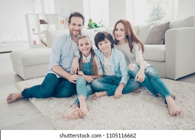 Portrait Of Lovely Cheerful Family With Two Kids In Casual Outfits Sitting On Carpet Near Sofa In Modern White Livingroom Looking At Camera. Domicile Domestic Lifestyle Rest Relax Leisure Concept