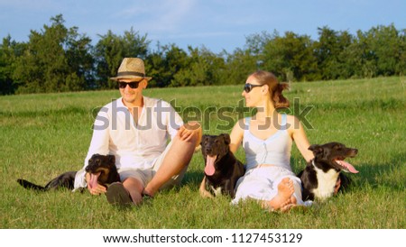 PORTRAIT: Lovely couple sits in a quiet grassy field with their three obedient black puppies. Lovely shot of boyfriend and girlfriend posing in a sunny meadow with their three cute border collies.