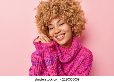 Portrait Of Lovely Curly Haired Young Woman Smiles Gently Keeps Hands Together Near Face Wears Knitted Winter Jumper Poses For Making Photo Looks Direcrly At Camera Isolated Over Pink Background