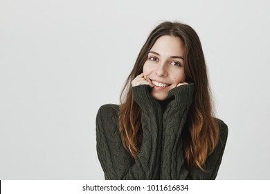 Portrait of lovely european female with dreamy look, holding hands on face, wearing green cozy sweater, isolated over white background. Girl smiles while talking to guy she likes - Shutterstock ID 1011616834