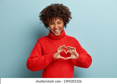 Portrait of lovely female model makes heart gesture, says be my valentine, demonstrates love sign, has glad expression, wears warm red jumper, isolated against blue background. Body language - Shutterstock ID 1395201326